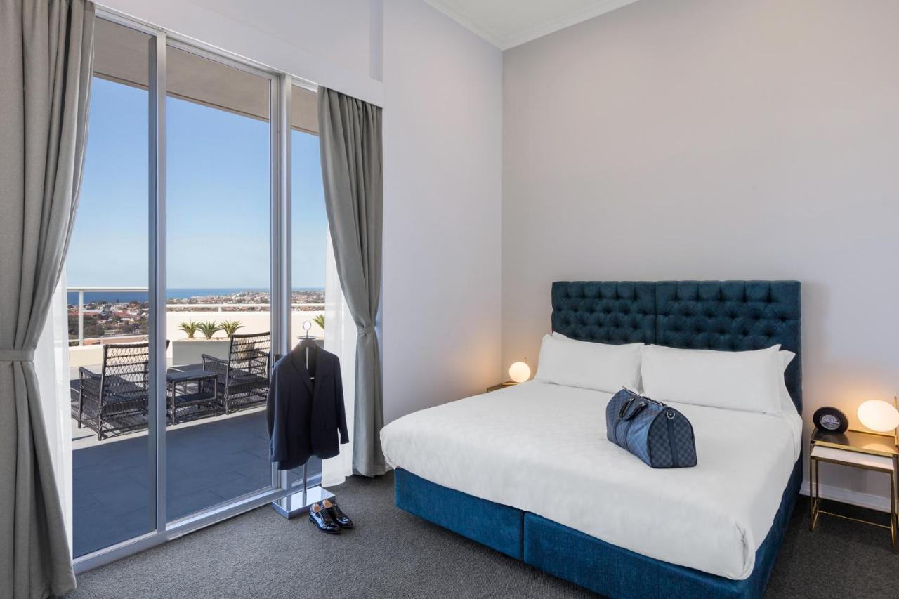 Meriton Suites Bondi Junction Sydney Luaran gambar The photo shows a stylish bedroom featuring a large bed with a tufted blue headboard. There's a neatly organized outfit consisting of a suit hanging on a stand, along with shoes placed on the floor. The room has large windows that provide a view of t