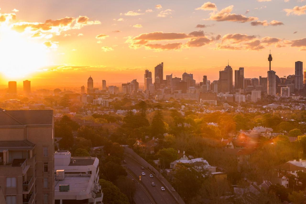 Meriton Suites Bondi Junction Sydney Luaran gambar The photo shows a panoramic view of a city skyline at sunset. The sun is low on the horizon, casting a warm golden light across the buildings. Tall skyscrapers are silhouetted against the colorful sky, which features hues of orange, pink, and yellow,