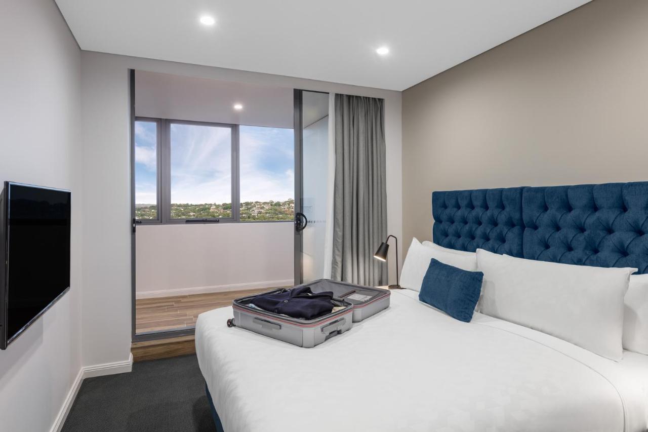 Meriton Suites Bondi Junction Sydney Luaran gambar The photo shows a modern hotel room featuring a double bed with a tufted blue headboard. There is a neatly arranged suitcase placed open on the bed, indicating that someone may be unpacking. The room has light-colored walls and a window allowing natu