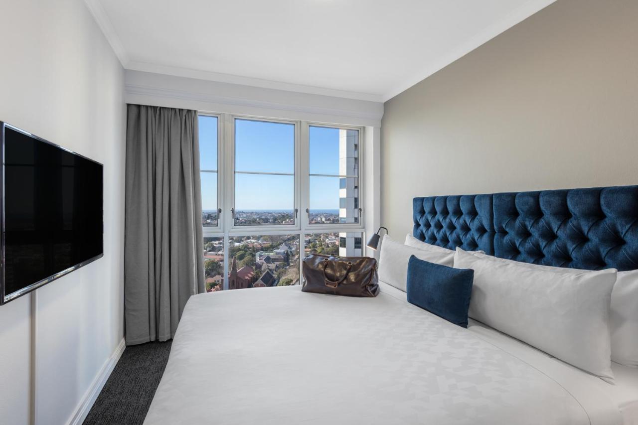Meriton Suites Bondi Junction Sydney Luaran gambar The photo shows a modern hotel room featuring a large bed with a tufted blue headboard. Beside the bed, there is a stylish bag placed on the bed. The room has large windows that provide a view of the outside, allowing natural light to fill the space.