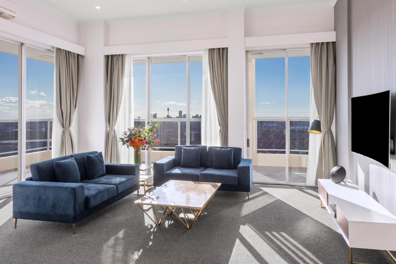 Meriton Suites Bondi Junction Sydney Luaran gambar The photo shows a modern living room with large windows that allow natural light to fill the space. There are two blue sofas arranged around a coffee table with a glass top and gold legs. A floral arrangement adds a touch of color, placed on a nearby
