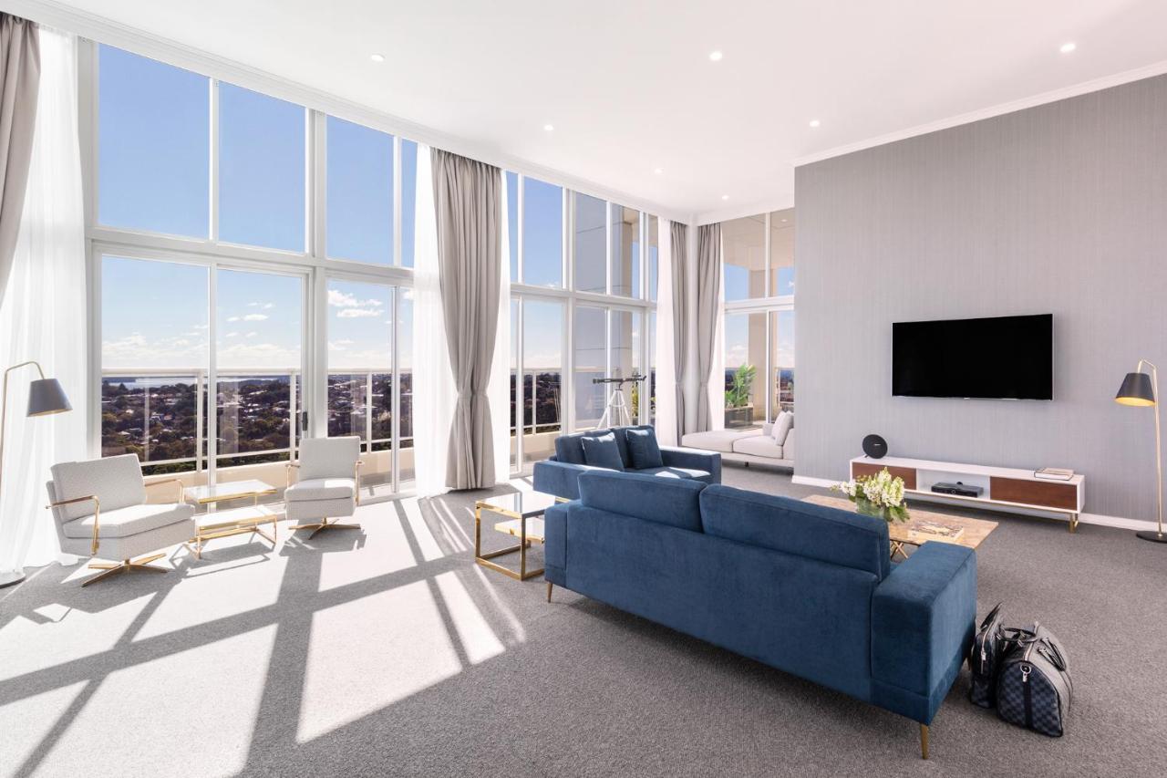 Meriton Suites Bondi Junction Sydney Luaran gambar The photo shows a bright and modern living room with large floor-to-ceiling windows that provide a view of the outdoors. The space features a blue sofa in the center, complemented by light-colored armchairs nearby. There are light curtains hanging by