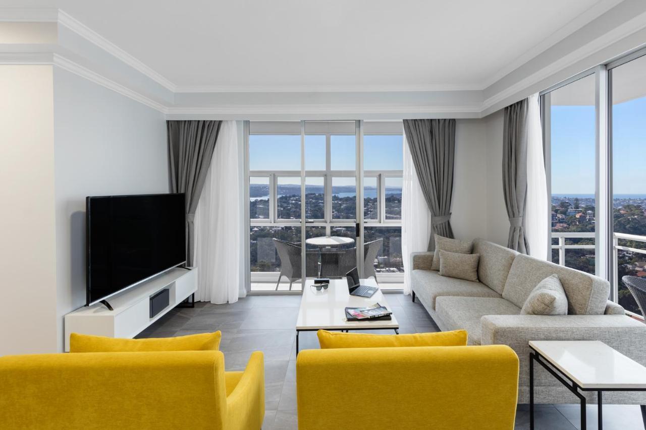 Meriton Suites Bondi Junction Sydney Luaran gambar The photo depicts a modern living room space with large windows that offer a view of the outside. The decor is minimalist and stylish, featuring a light color palette. 

In the center, there are two bright yellow chairs facing each other. A sleek, re