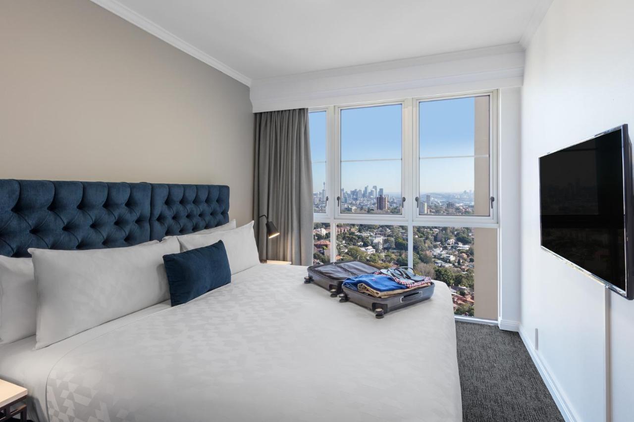 Meriton Suites Bondi Junction Sydney Luaran gambar The photo shows a modern hotel bedroom featuring a king-sized bed with a blue tufted headboard. The bed is neatly made with white linens and a decorative blue pillow. There is a large window that offers a view of the city skyline, allowing natural li