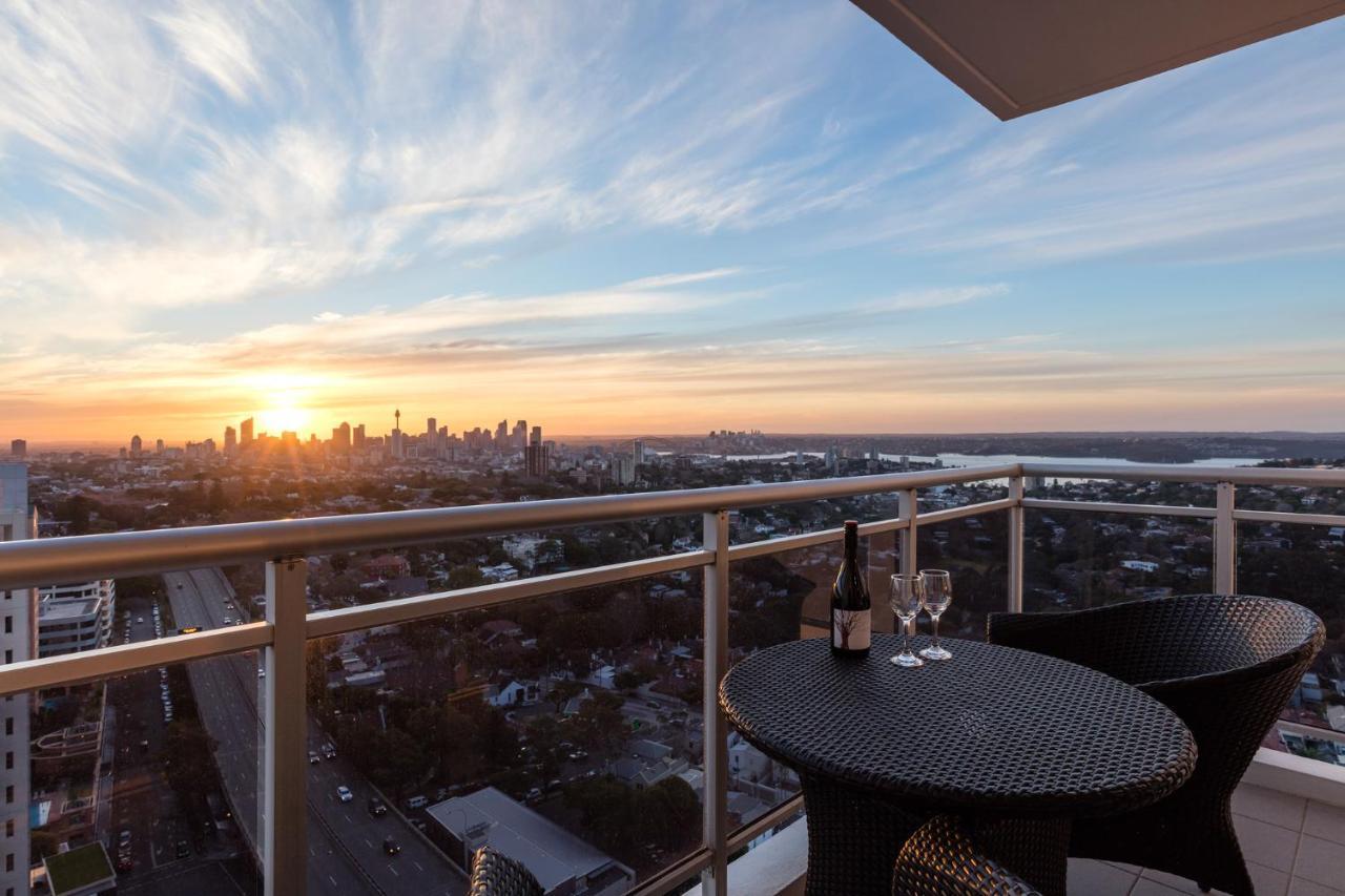 Meriton Suites Bondi Junction Sydney Luaran gambar The image shows a scenic view from a balcony or terrace overlooking a cityscape during sunset. In the foreground, there is a small round table with two glasses and a bottle of wine, suggesting a romantic or relaxing setting. The sky is filled with vi
