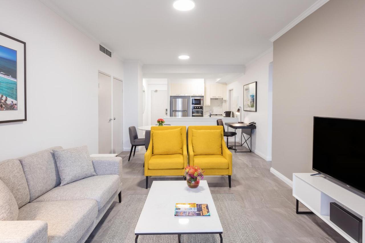 Meriton Suites Bondi Junction Sydney Luaran gambar The photo shows a modern living space with a light and airy design. In the foreground, there are two bright yellow armchairs flanking a white coffee table, which has a small vase of flowers and a magazine on it. To the left, there is a light gray sof