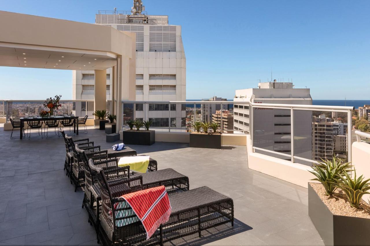 Meriton Suites Bondi Junction Sydney Luaran gambar The photo shows a rooftop terrace with a modern design. There are several outdoor seating arrangements featuring black wicker lounge chairs. Some of the chairs have colorful blankets draped over them. The terrace is surrounded by a railing and has po