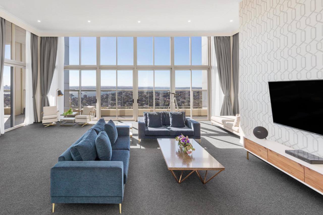 Meriton Suites Bondi Junction Sydney Luaran gambar The photo shows a modern and spacious living area. It features large windows that provide a panoramic view of the outside landscape and allow natural light to fill the room. The interior design includes a blue sectional sofa placed in the center, com