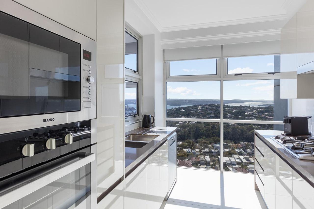 Meriton Suites Bondi Junction Sydney Luaran gambar The photo shows a modern kitchen with sleek, shiny white cabinets and a large window that offers a panoramic view of the outside landscape. The kitchen features built-in appliances, including an oven and a stove, with a stylish countertop. The spacio