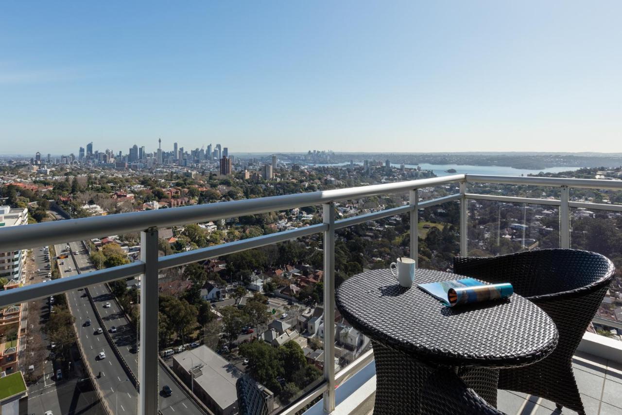 Meriton Suites Bondi Junction Sydney Luaran gambar The photo shows a balcony with a view overlooking a cityscape and a body of water in the distance. There is a round table with a woven design and two chairs, one of which has a mug on it. On the table, there’s also a rolled-up magazine or document. T