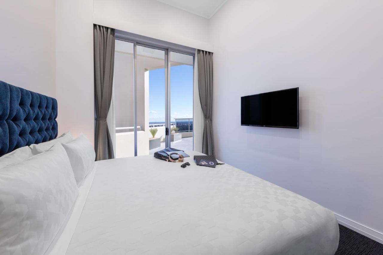 Meriton Suites Bondi Junction Sydney Luaran gambar The photo shows a modern hotel room with a large, comfortable bed featuring a blue tufted headboard. There are light-colored walls, and the room is decorated simply. A flat-screen TV is mounted on the wall opposite the bed. The room has large glass d