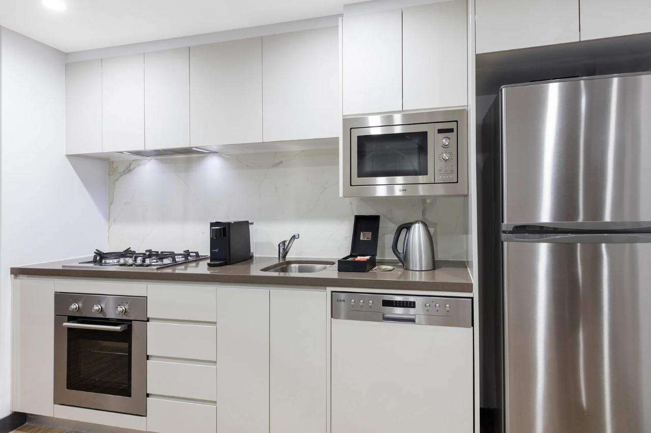 Meriton Suites Bondi Junction Sydney Luaran gambar The photo shows a modern kitchen with a sleek, minimalist design. It features white cabinetry and a countertop that blends with the overall aesthetic. 

Key elements include:

- A stainless steel refrigerator on the right.
- An oven with a gas cookto