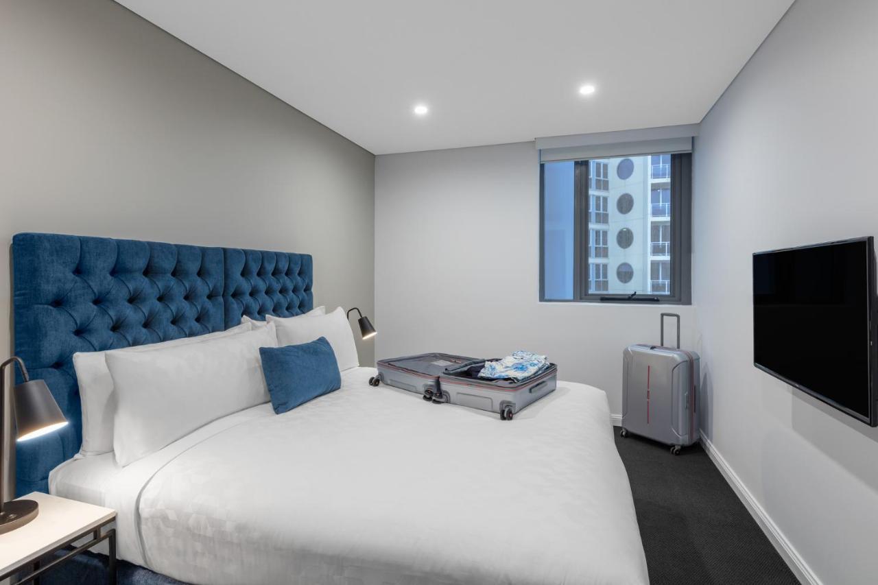 Meriton Suites Bondi Junction Sydney Luaran gambar The photo shows a modern hotel room featuring a neatly made bed with a blue tufted headboard. There is a small blue decorative pillow on the bed, and the bedding is white. Next to the bed, there is a nightstand with a lamp. A suitcase is partially op