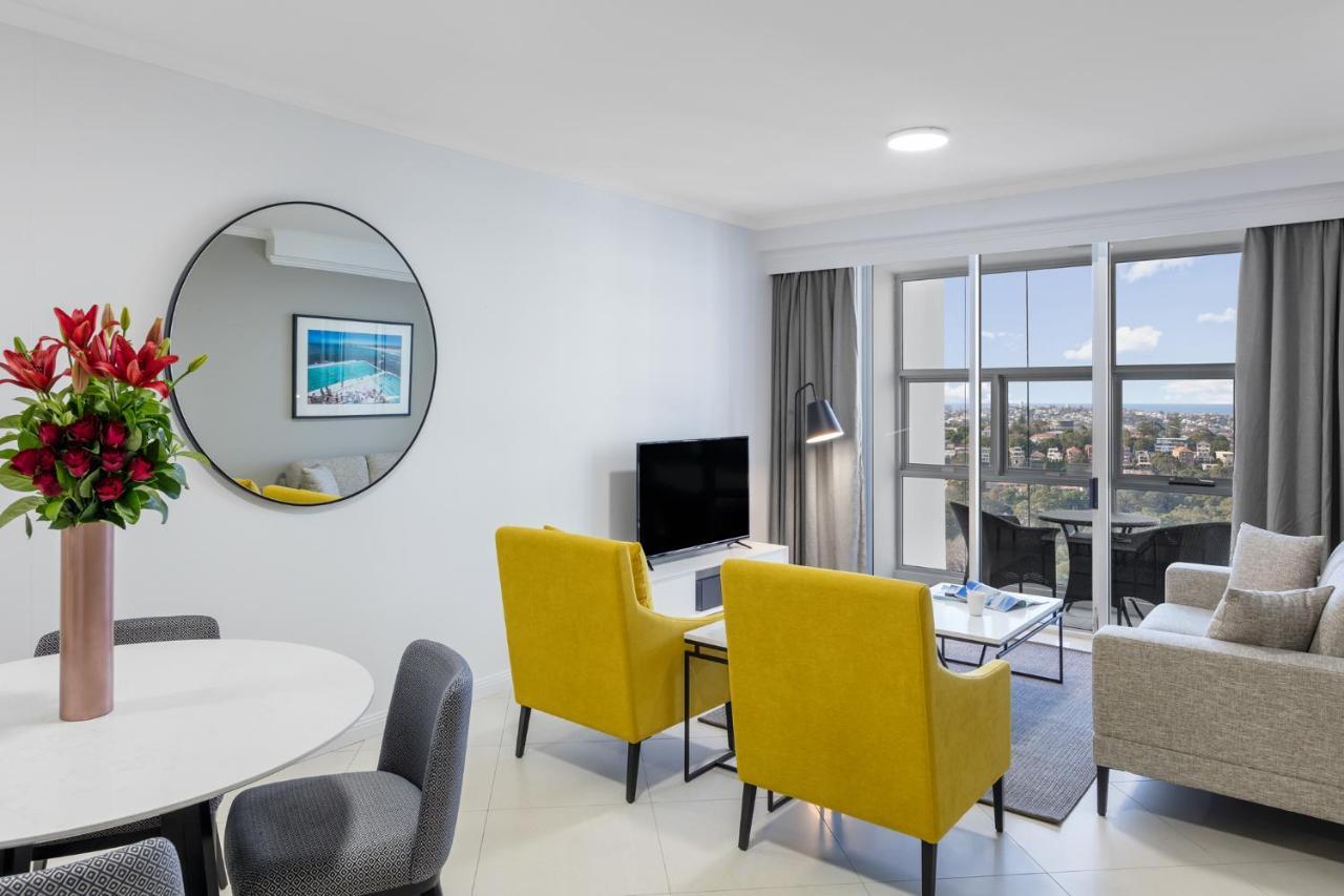 Meriton Suites Bondi Junction Sydney Luaran gambar The photo shows a modern living space with a stylish design. There are two bright yellow chairs facing a small glass table, and a comfortable beige sofa to the right. A TV is positioned on the wall in front of the chairs. To the left, there's a dinin