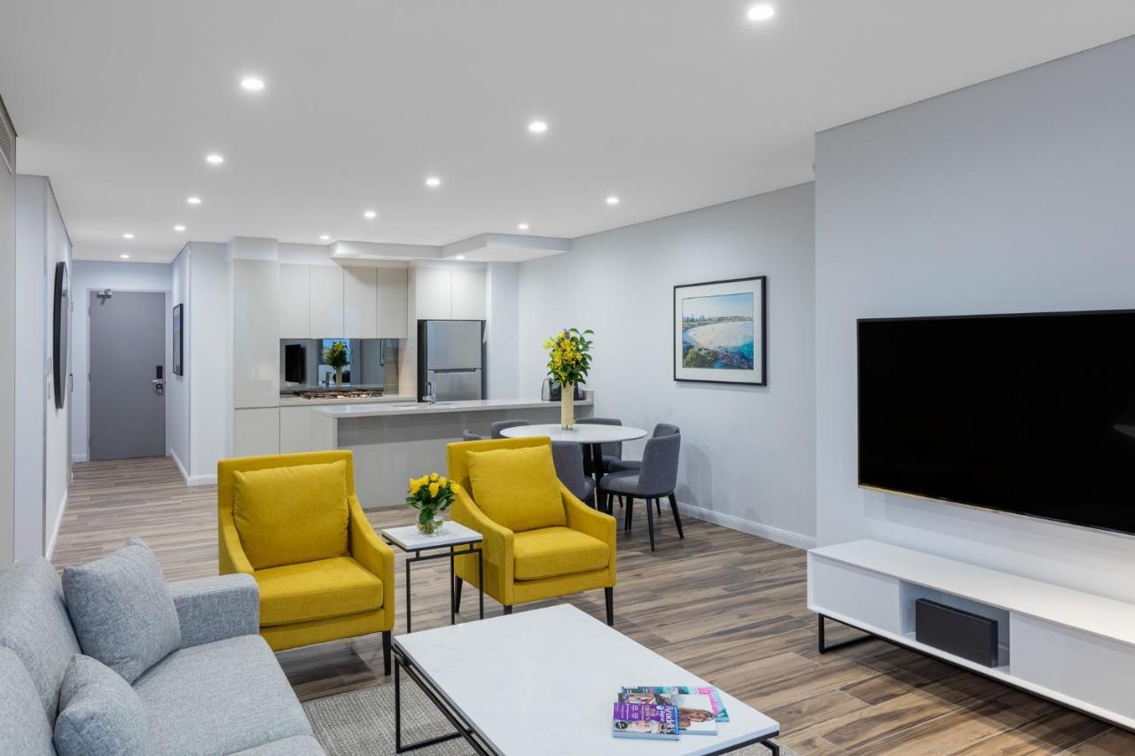 Meriton Suites Bondi Junction Sydney Luaran gambar The photo shows a modern living space that includes a cozy sitting area and a dining area. The sitting area features two bright yellow chairs and a light gray sofa, arranged around a coffee table. There are decorative items on the table, including fl
