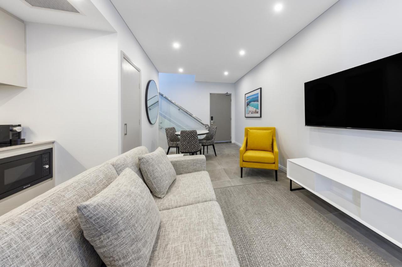 Meriton Suites Bondi Junction Sydney Luaran gambar The photo shows a modern and stylish living space. It features a light-colored sofa with cushions, positioned to face a wall-mounted flat-screen TV. There's a yellow accent chair nearby that adds a pop of color to the room. The décor is minimalistic,