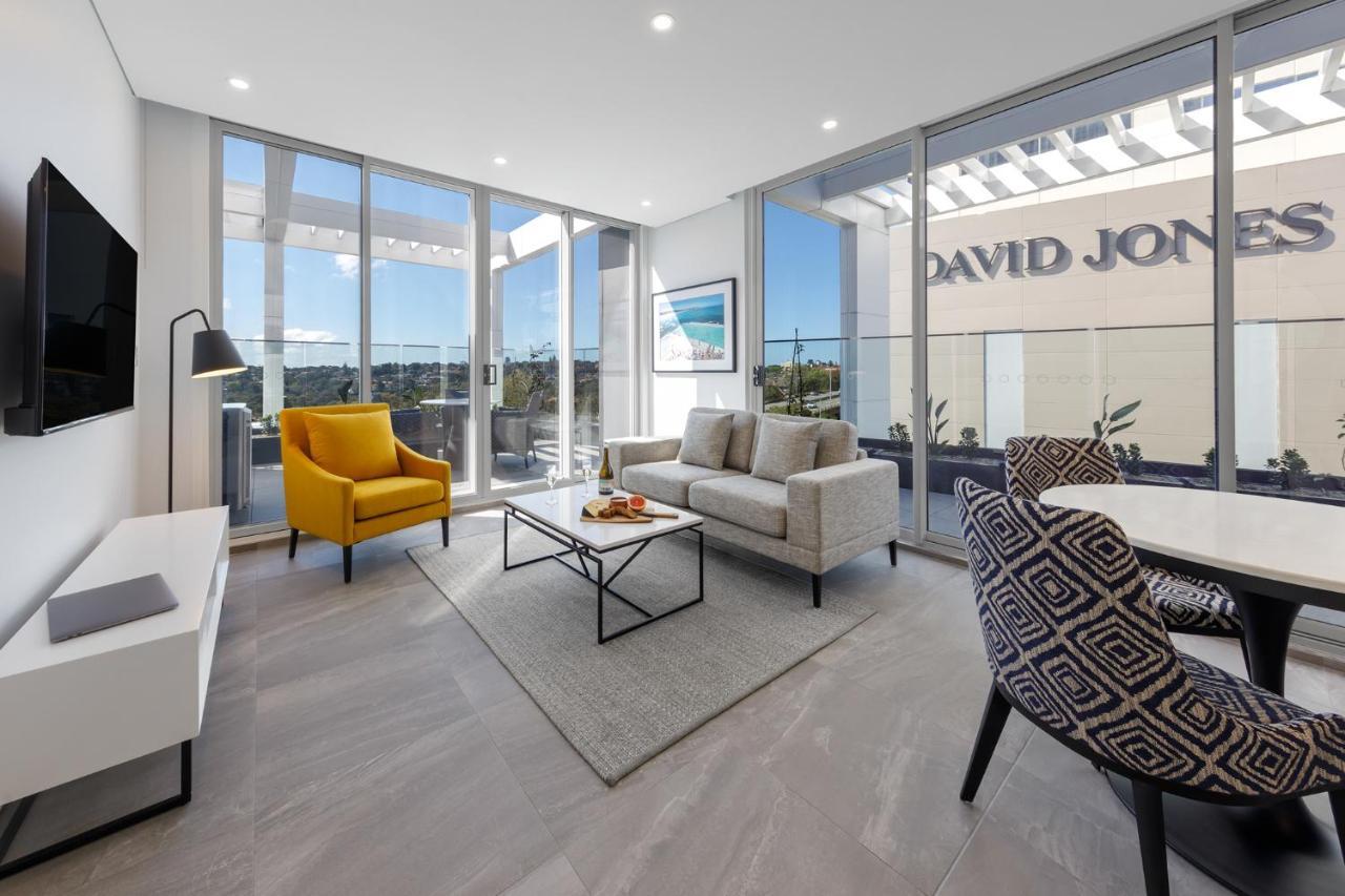 Meriton Suites Bondi Junction Sydney Luaran gambar The photo shows a modern living space that features large windows allowing ample natural light. The room is furnished with a gray sofa and a bright yellow armchair, along with a coffee table in between. There’s a decorative bowl with fruits on the ta