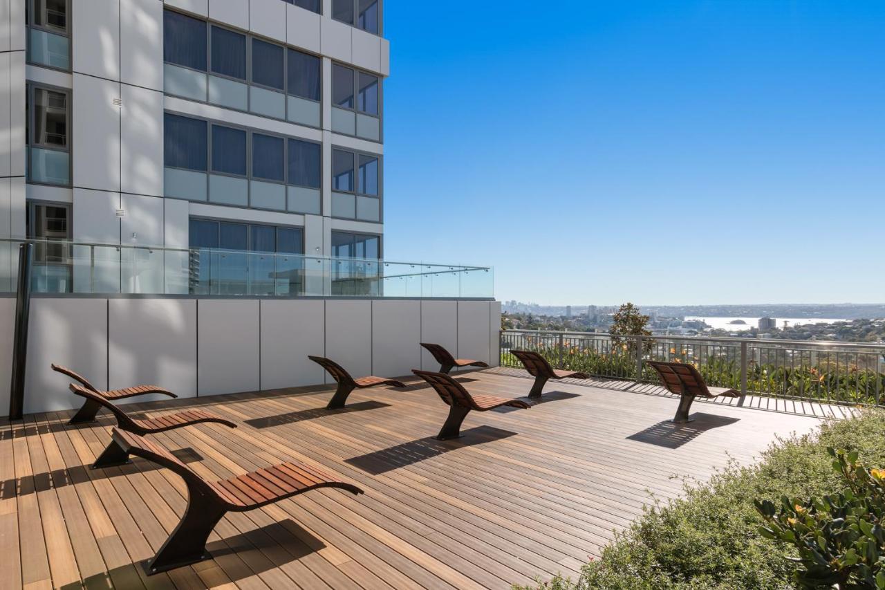 Meriton Suites Bondi Junction Sydney Luaran gambar The photo shows a rooftop or outdoor area with a wooden deck. There are several modern lounge chairs arranged neatly, facing a clear sky and a panoramic view of the surrounding landscape. In the background, there is a tall building with large windows