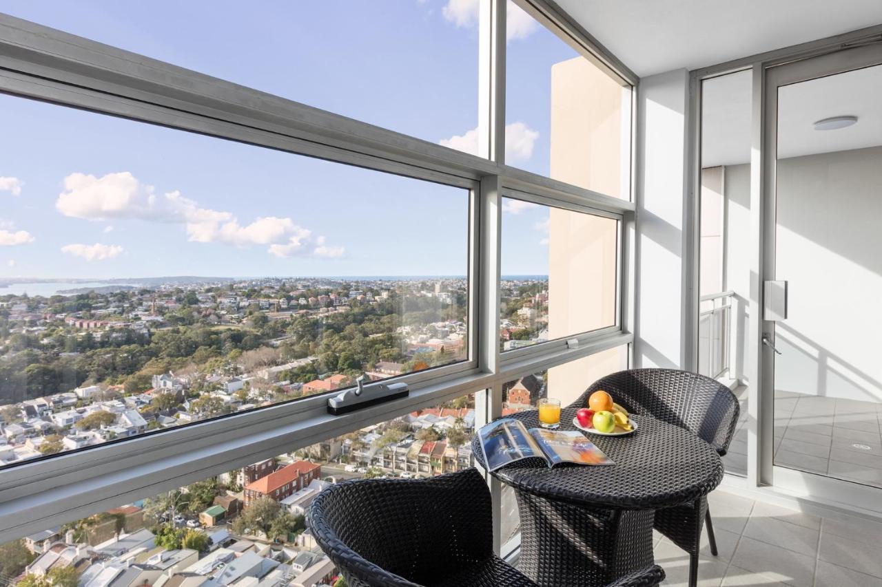 Meriton Suites Bondi Junction Sydney Luaran gambar The photo shows a modern balcony with large glass windows that offer a panoramic view of a sprawling urban landscape. There is a small round table made of dark wicker, accompanied by two matching chairs. On the table, there is a bowl of fruit and a m