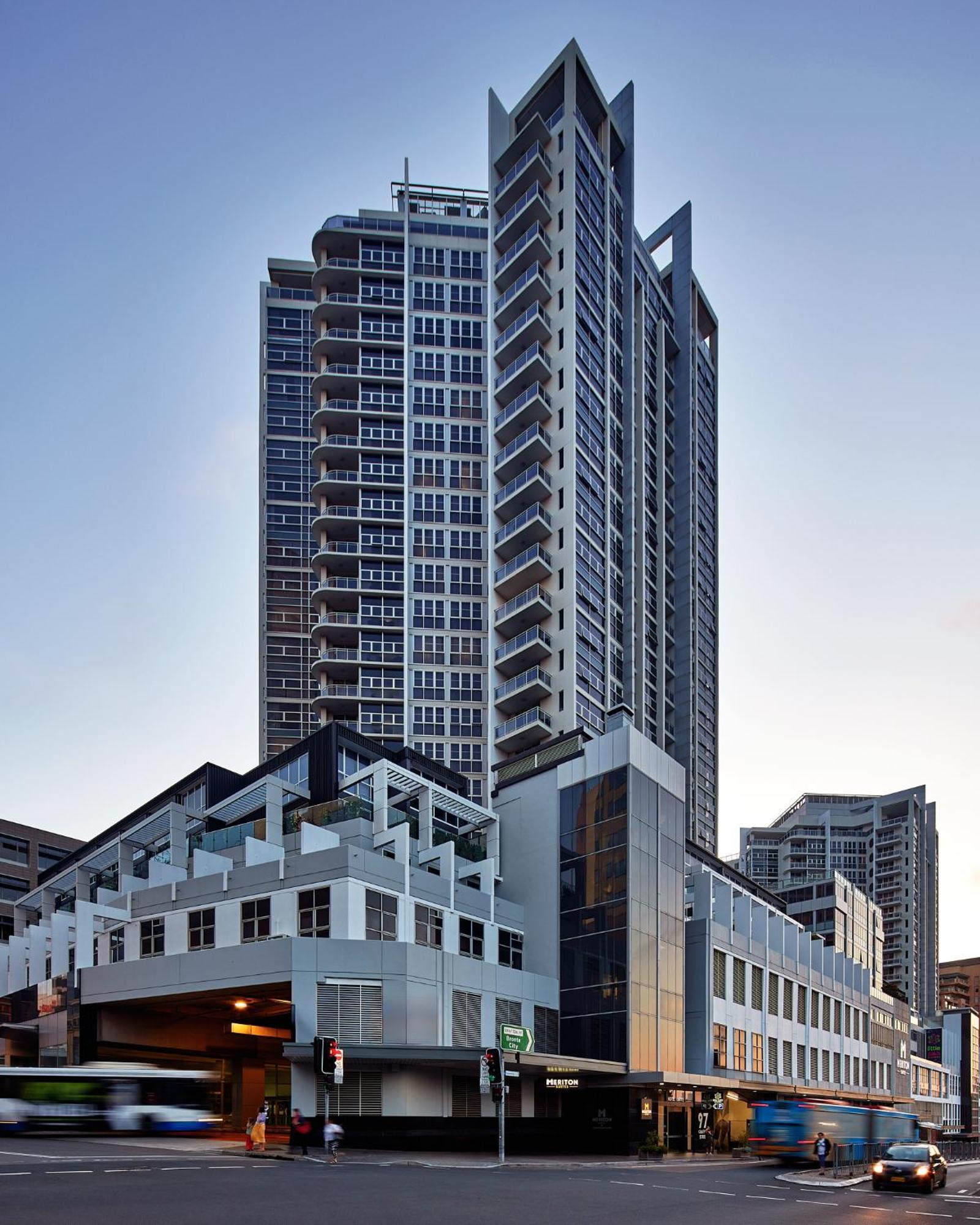 Meriton Suites Bondi Junction Sydney Luaran gambar The image shows a modern high-rise building with a sleek design. The structure features a mix of glass and concrete elements, with multiple levels showcasing balconies. The building appears to be part of an urban environment, likely containing reside
