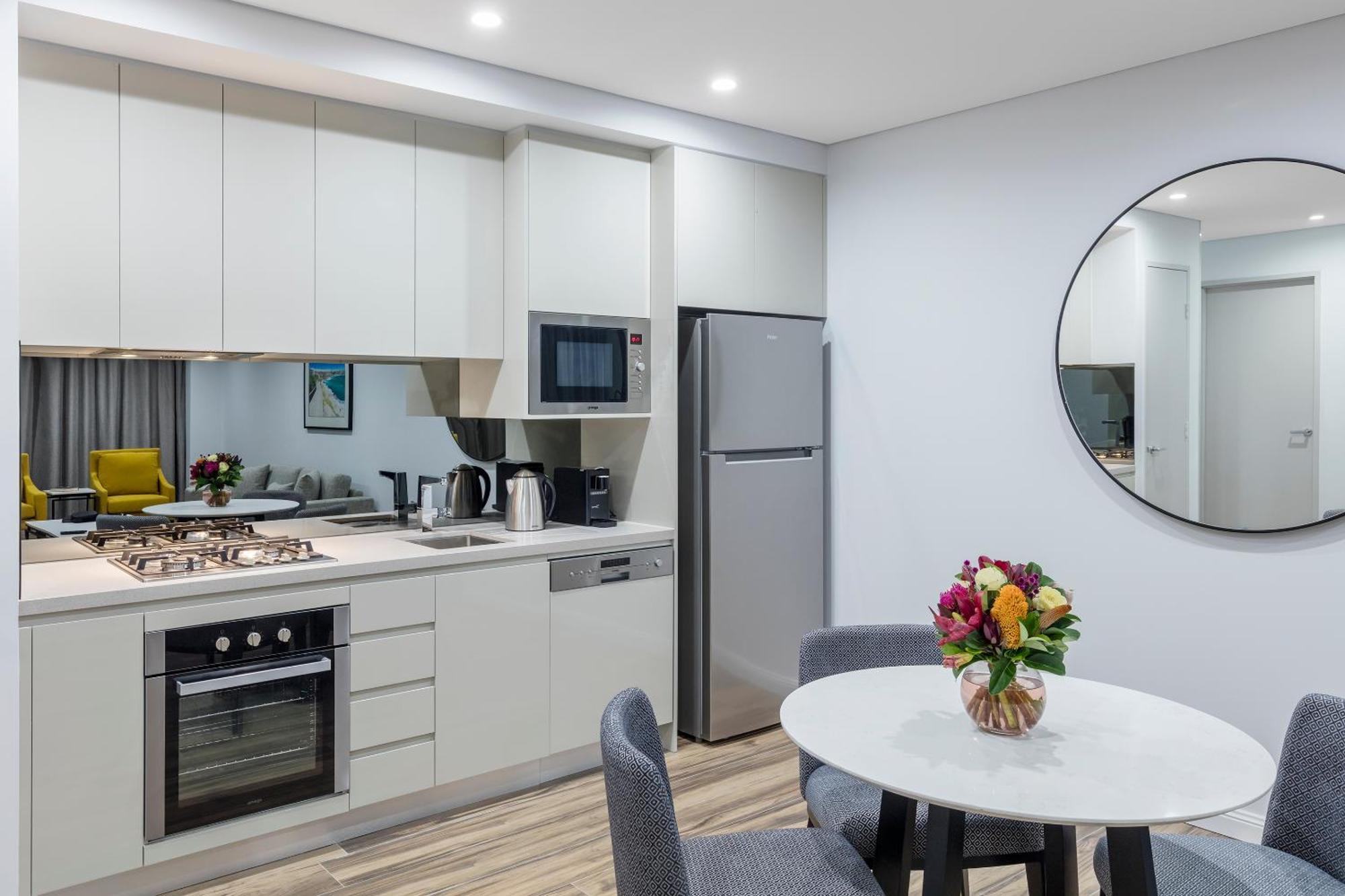 Meriton Suites Bondi Junction Sydney Luaran gambar The photo shows a modern kitchen and dining area. The kitchen features light-colored cabinetry with a built-in oven and stovetop, a microwave, and a silver refrigerator. There is a kettle and a coffee maker on the countertop. In the dining area, ther