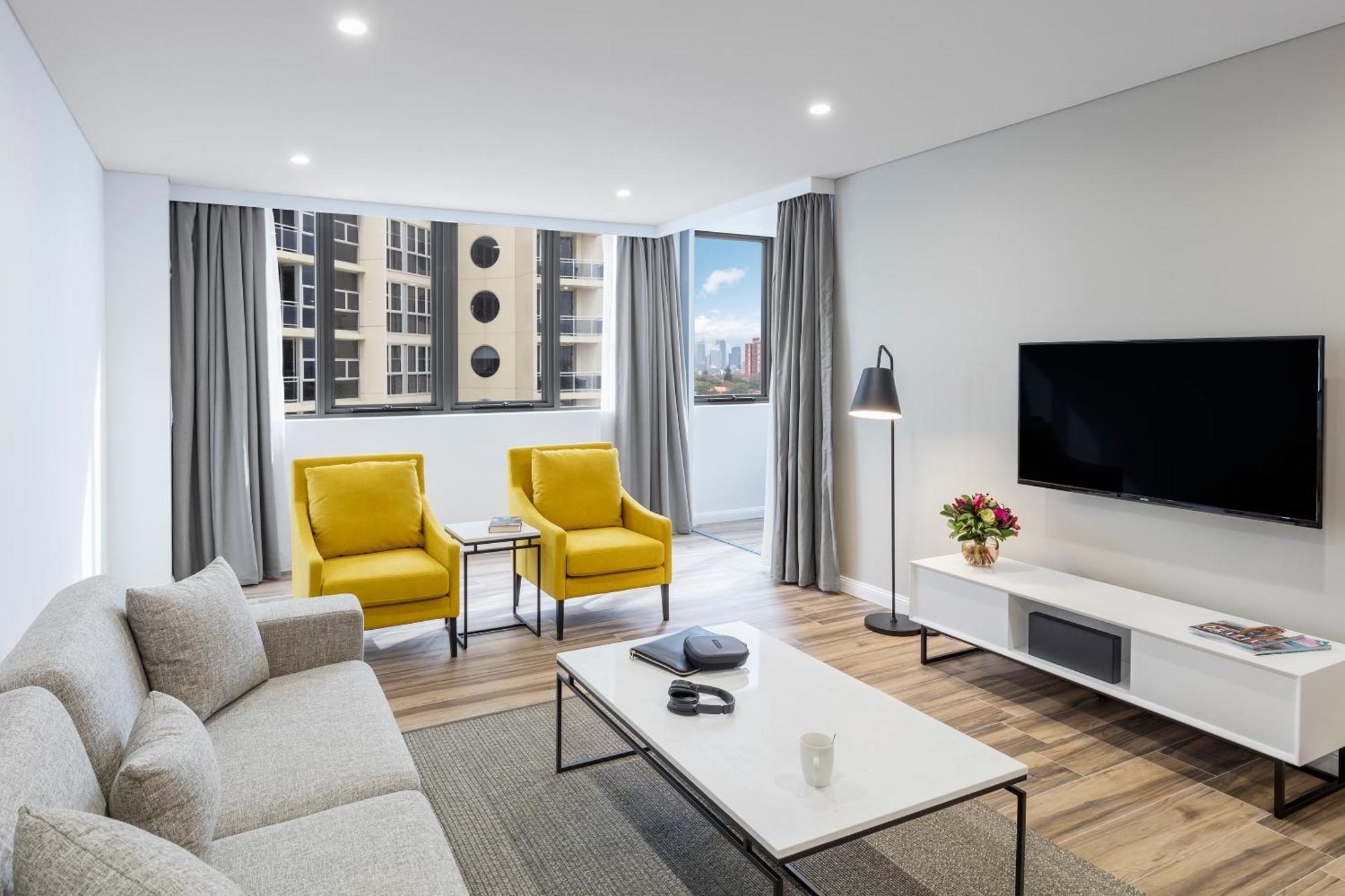 Meriton Suites Bondi Junction Sydney Luaran gambar The photo shows a modern, well-lit living room. It features a light gray sofa with a textured fabric. There are two bright yellow armchairs positioned near a window, which allows natural light to enter the space. The room has light gray curtains and 